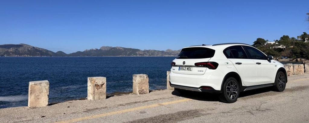 Weißer Fiat Tipo Cross Hybrid mit spanischem Kennzeichen parkt an einer Küstenstraße auf Mallorca mit Blick auf das Mittelmeer und die Tramuntana-Berge im Hintergrund an einem sonnigen Tag.