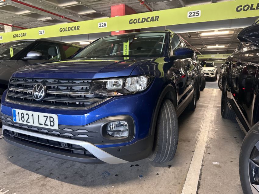 Ein blauer VW T-Cross mit spanischem Kennzeichen steht auf einem markierten Parkplatz im Parkhaus der Autovermietung Goldcar. Weitere Fahrzeuge sind in den angrenzenden Stellplätzen sichtbar.
