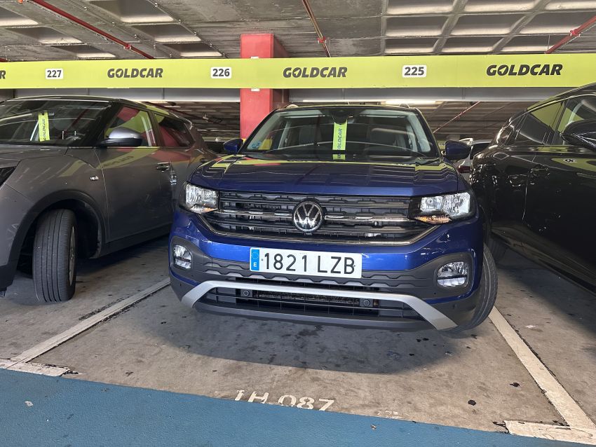 Ein blauer VW T-Cross mit spanischem Kennzeichen 1821 LZB steht auf einem Goldcar-Parkplatz in einem überdachten Parkhaus.